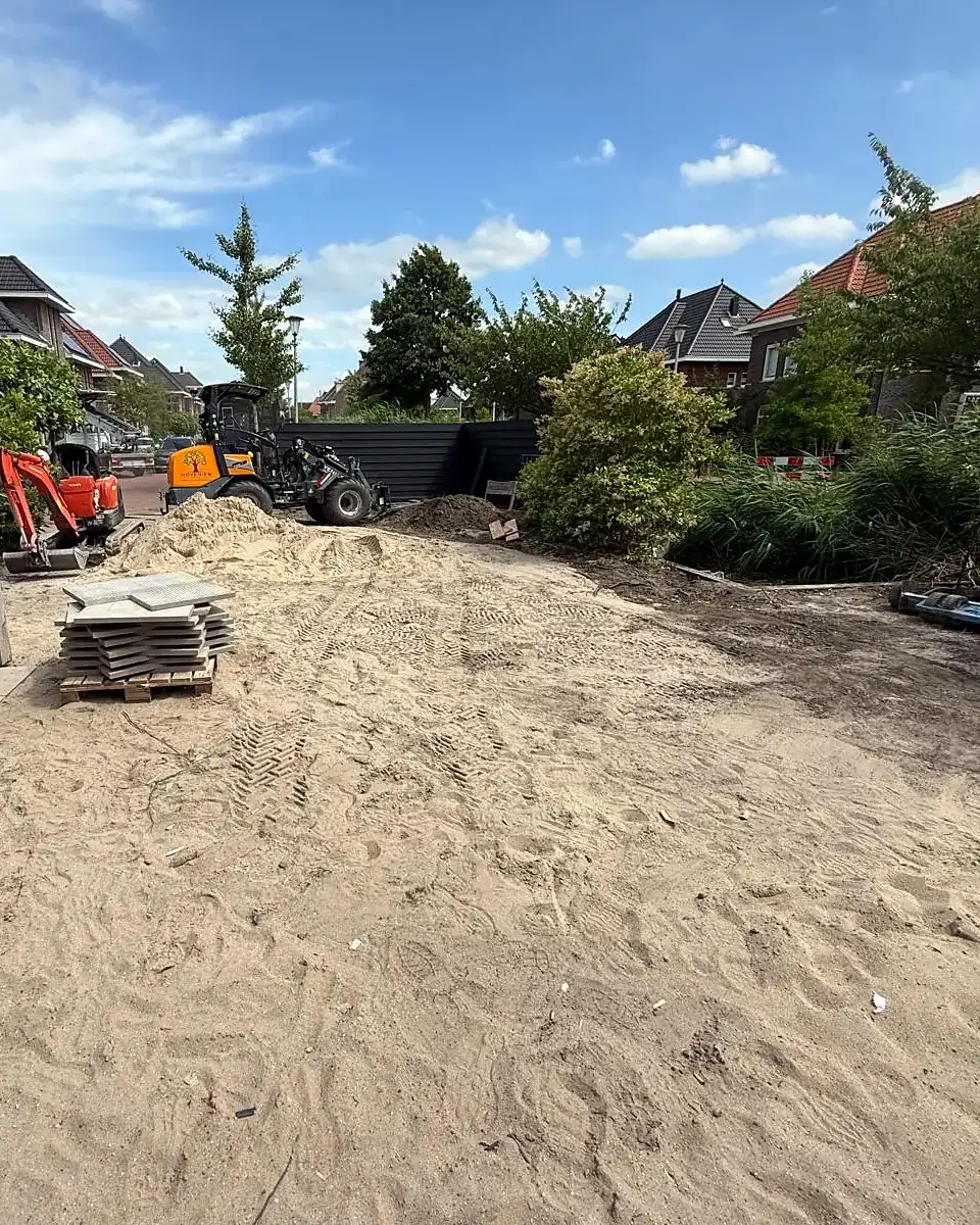 Vóór: zandvlakte met stapels tegels en het materieel achter in de tuin